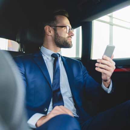 Confident successful rich young caucasian businessman in formal clothes using smart phone while sitting at backseat of taxi car with a driver looking at the window.